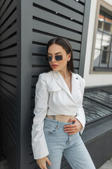 Fashionable young beautiful woman with vintage sunglasses in white fashion top blazer and blue jeans stands near a black wall on the street