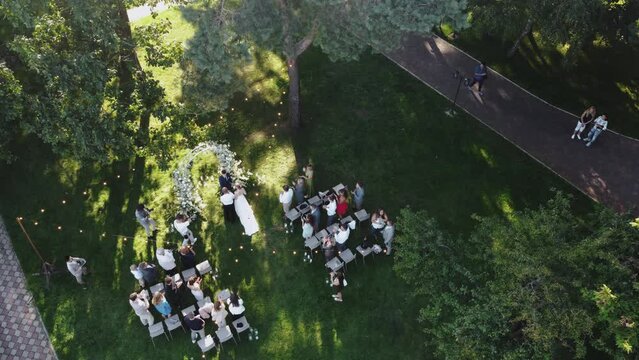 Aerial top view of wedding ceremony in the park with arch decorated with white flowers and the the guests stand and applaud the bride as she goes to the groom , unrecognisable people. 