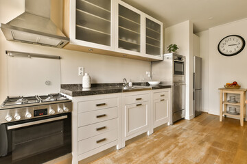 a kitchen with wood flooring and white cupboards on the wall behind it is a large clock hanging above the stove