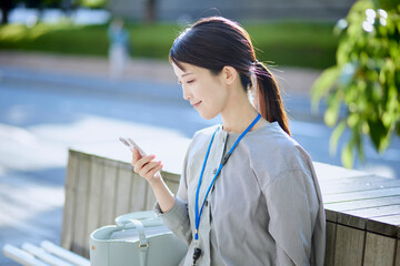 仕事の休憩中にベンチに座りスマートフォンを操作する日本人女性 - obrazy, fototapety, plakaty