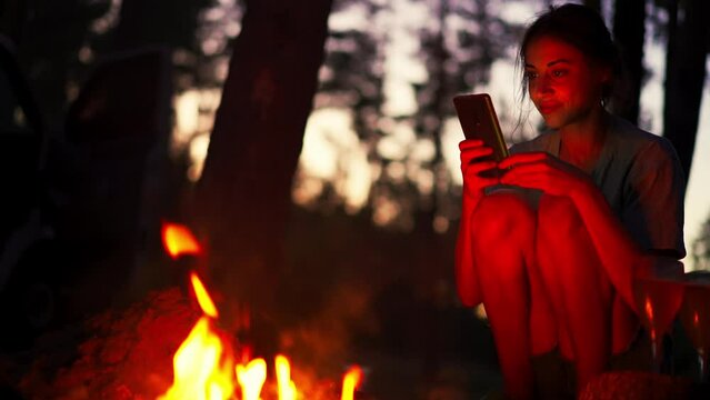 Hipster girl using smartphone sitting next to bonfire in forest camping at night. Woman hiking alone. Active leisure and lifestyle. People in woods. Tourist woman in forest campsite.