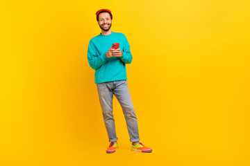 Fototapeta na wymiar Full size photo of optimistic guy wear teal sweatshirt red hat hold smartphone in hands read email isolated on yellow color background