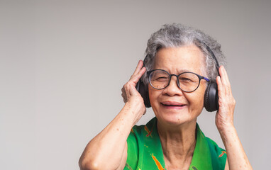 Cheerful senior woman wearing wireless headphones and hands up with a smile while standing on a gray background.