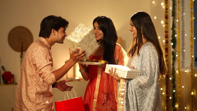 Indian man giving gift to wife and sister during diwali festivel and wife feeding sweet to husband.they all are very happy.well decorated home,hindu festival