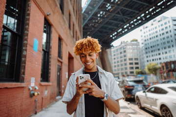 Young handsome guy walks across the street, typing messages on a smartphone and smiling
