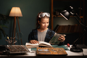Schoolgirl soldering computer circuit with soldering iron and reading book in her room at home