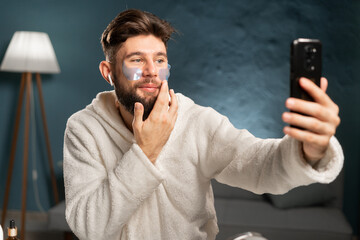 Man with collagen patches under eyes after bath in bathrobe shooting selfie on front camera...