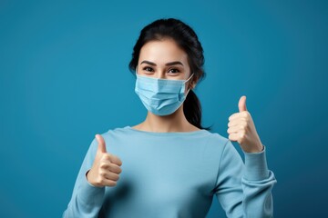 young woman in medical mask giving thumbs up gesture on blue background. - obrazy, fototapety, plakaty