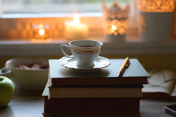 Vintage books, e-reader, pen, reading glasses, phone, cup of tea or coffee, bowl of cookies, green apple and lit candles on the table. Hygge at home, studying atmosphere. Selective focus.