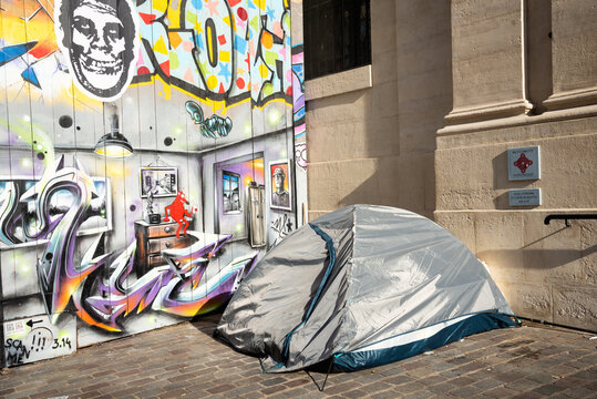 Paris, France - February 21, 2023: Social contrasts. Tent of homeless person at street, apartment graffiti on house wall and plate of Historic Monument (belonging to a church beside). 