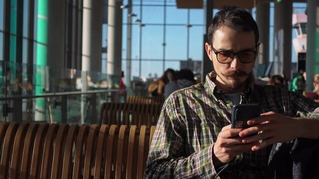 Young adult Caucasian man sits in departure hall of Dalaman airport with smartphone in hands and waits. He is wearing casual clothes and looking happy because of coming holiday. Travel to Turkey theme
