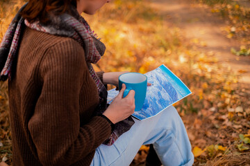 woman with a backpack drinks tea from a cup, dressed in a brown sweater, holds a map in her hands, enjoys sunny weather in the autumn forest, traveler on the precipice, hiking - obrazy, fototapety, plakaty