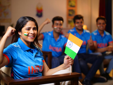 Bokeh shot of cricket fans watching a live match at home - a female fan holding an Indian flag in her hands  cheering up for team India. A group of friends huddled together- wearing a jersey  cheer...
