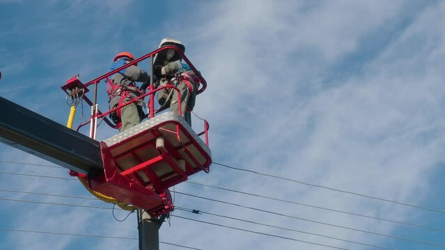 Two professional electricians from cab of aerial platform are repairing street lighting lamp. Video of two electricians working at their best. View from bottom to top against blue sky.