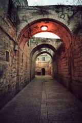 Fototapeta na wymiar Ancient deserted alley in Jewish Quarter in the Old City Jerusalem with switched on street light on the vault. Old narrow street with archeway construction. Empty road of old quarter in early morning.