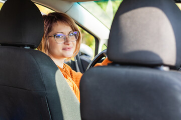 Happy owner in his newly purchased car sits relaxed, smiling happily. Beautiful young adult 30-35 years old woman in eyeglasses. Getting a driver's license, driving and having fun, learning to drive - obrazy, fototapety, plakaty