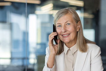 Close-up portrait of a smiling senior business woman in a suit sitting at a desk in the office and...