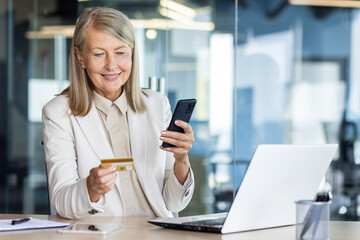 Senior smiling gray-haired business woman in a suit is working in the office on a laptop. Holds a phone and uses a credit card through an online application.