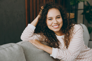 Close up young smiling satisfied minded woman wear casual clothes sits on grey sofa couch looking camera stay at home hotel flat rest relax spend free spare time in living room indoor. Lounge concept.