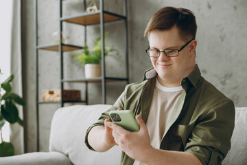 Young man with down syndrome wear glasses casual clothes use mobile cell phone chatting sits on grey sofa couch stay at home flat rest spend free time in living room Genetic disease world day concept - obrazy, fototapety, plakaty