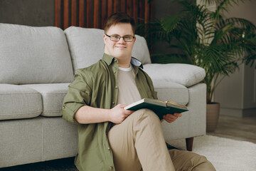 Side view young smart man with down syndrome wearing glasses casual clothes reading book sit near...