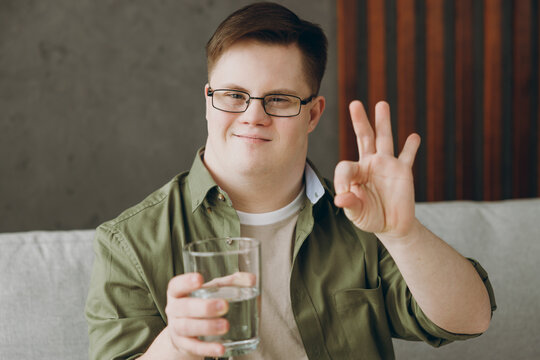 Young man with down syndrome in glasses casual clothes drink water show ok okay gesture sit on grey sofa couch stay at home flat rest spend free time in living room Genetic disease world day concept