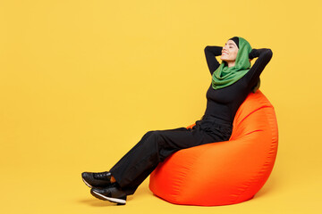 Full body young arabian asian muslim woman wears green hijab abaya black clothes sit in bag chair hold hands behind neck rest isolated on plain yellow background. People uae islam religious concept.