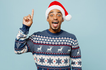 Young smart fun man wear knitted sweater Santa hat posing holding index finger up with great new...
