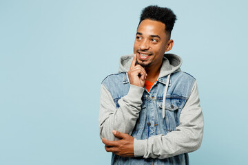 Young man of African American ethnicity wearing denim jacket orange t-shirt put hand proping up on chin, lost in thought and conjectures isolated on plain pastel light blue background studio portrait. - obrazy, fototapety, plakaty