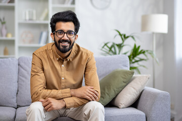 Portrait of young idian man in glasses and shirt sitting on sofa at home in front of camera and smiling