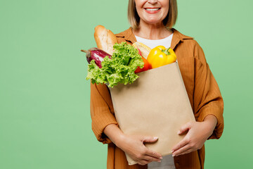Close up cropped elderly smiling happy woman wear brown shirt casual clothes hold shopping paper bag with food products looking camera isolated on plain green background. Delivery service from shop.