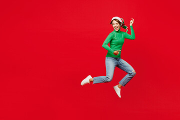 Full body side view young woman in green turtleneck Santa hat posing jump high pov play guitar hand gesture isolated on plain red background Happy New Year 2024 celebration Christmas holiday concept