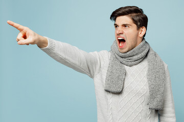 Young ill sick man wears gray sweater scarf point index finger aside scream isolated on plain blue...