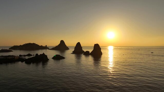 Gabbiano vola all'alba tra i Faraglioni dei Ciclopi di Aci Trezza, Sicilia, Italia.
Ripresa aerea controluce degli scogli sulla costa siciliana con il sole giallo delle prime luci dell'alba. 