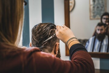 Hipster man at barbershop salon getting beard and hair cut - Hairdresser woman using hair scissors and comb for to modern gentleman cut - Barber shop concept - obrazy, fototapety, plakaty