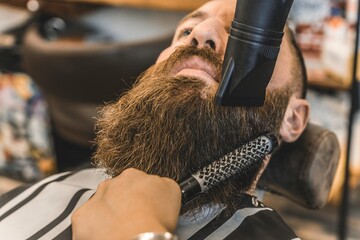 Female hairdresser using the hair dryer to straighten his long beard - Hipster man at barbershop salon getting beard and hair cut - Barber shop concept - obrazy, fototapety, plakaty