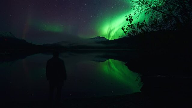 Moment of reflection beneath an aurora in October. Silhouette of a man gazing across a silent lake, in a mountainous landscape beneath beautiful auroras.