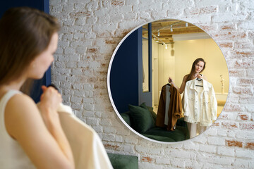 A young woman looks at herself in a round mirror and chooses between clothes to wear outside. The...