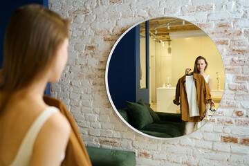 A young woman looks at herself in a round mirror and chooses between clothes to wear outside. The concept of changing the wardrobe, buying new things and styling