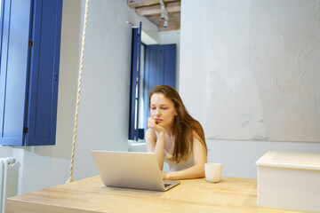 Young woman thoughtfully looking at laptop while sitting at wooden table in room at home. The...