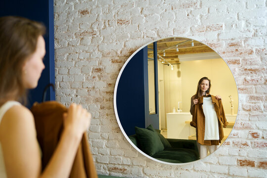 A young woman is trying on clothes on a hanger while looking at a round mirror in a room. The concept of changing the wardrobe, styling and choosing a look