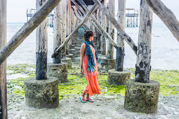 Youg beautiful woman posing for camera near wooden sea dock pillars in the water stretching into the sea, Nungwi, Zanzibar, Tanzania