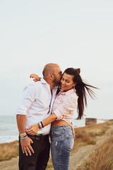 Happy smiling  Couple walking at Sand Dunes near the Beach.  Young happy Bearded muscular  man  in White shirt kissing and hugging beautiful woman at sunset on a beach
