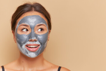 Cute young woman with clay mask on her face posing over background with gentle smile, skincare routine concept, copy space