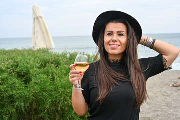 Fototapeta premium Portrait of Young Beautiful Smiling happy woman standing with glass of wine against sea during the winter in leather jacket and hat .