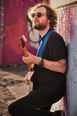 Hipster street musician in black playing electric guitar in the street on sunset leaning on a wall