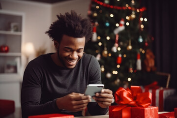 Young afro american male ordering a christmas present on phone sitting next to a christmas tree. Smiling happy young african male holding phone.