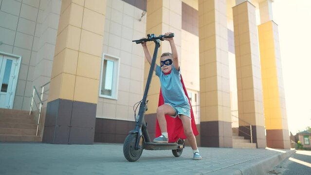 Girl superhero on electric scooter. happy family kid concept. a child on electric scooter rides on a large square in the city center under a blue sky with clouds and rays of dream sun. superhero girl