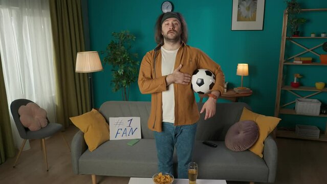 In the picture, a middle-aged man stands near the sofa. Putting his hand on his heart, the second holds the ball. Shows watching a football match. Then he kisses the ball and sits in a choke hold