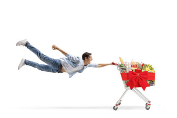 Full length shot of a casual young man flying and holding a shopping cart with food and red ribbon...
